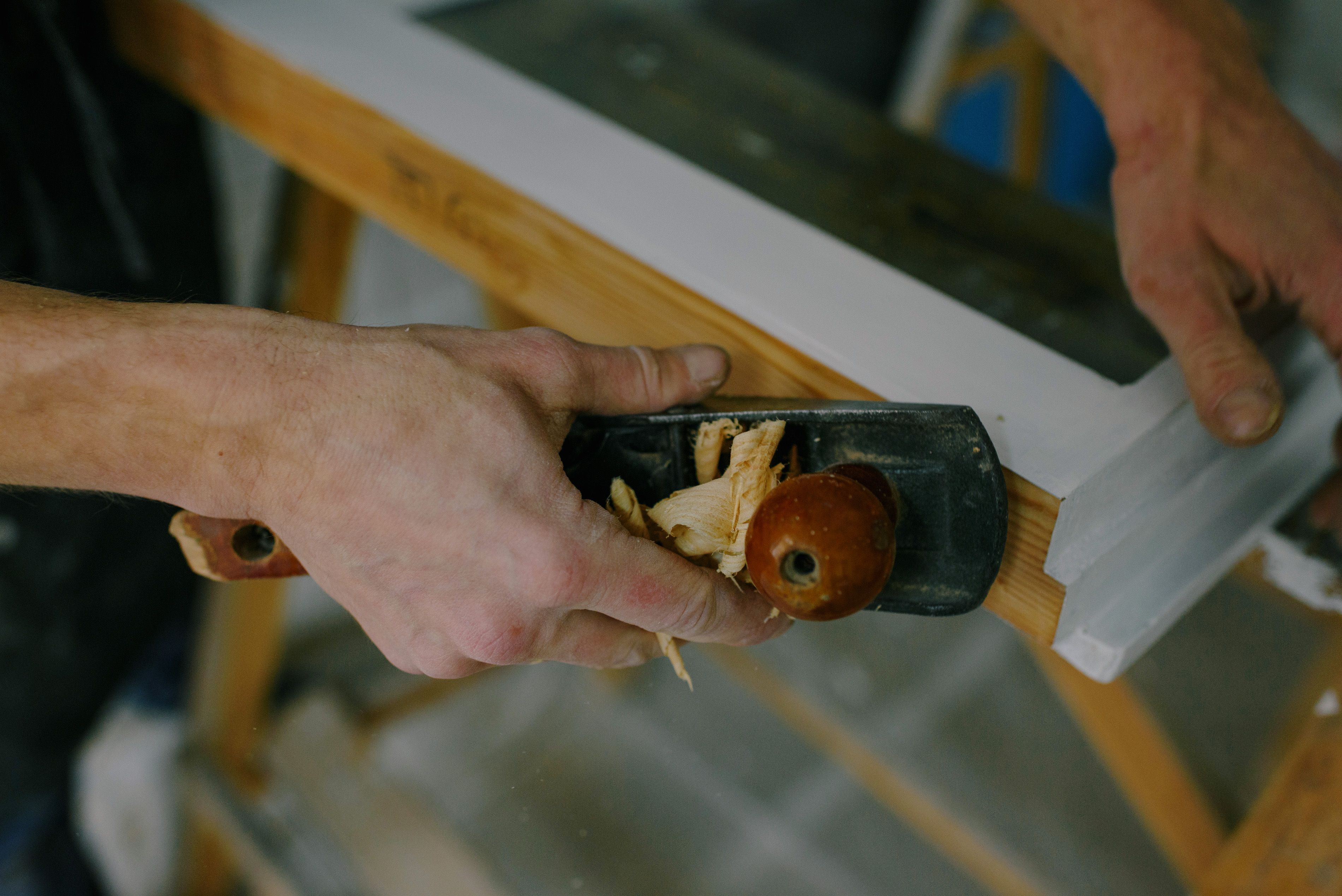 Close-up of hands using a hand plane on a piece of white-painted trim, with wood shavings curling up from the blade